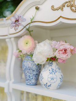 Two blue and white floral vases, one taller with a ranunculus and the other rounder with a peony and roses, sit on a white, ornate shelf.