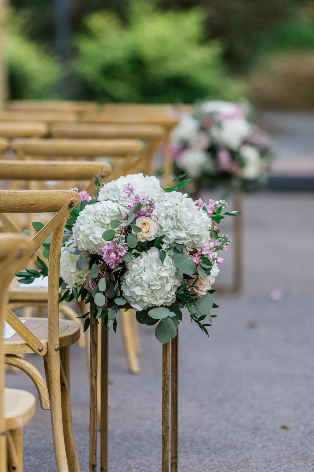 close up on vineyard chairs WEBP Light colored X-back chairs in rows at an outdoor wedding ceremony. Florals on the ends of each row.