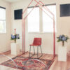 Copper arch with 3 peaks against a white wall with windows set up for a ceremony. Rug in front and florals on sides.