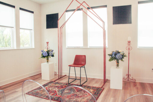 Copper arch with 3 peaks against a white wall with windows set up for a ceremony. Rug in front and florals on sides.