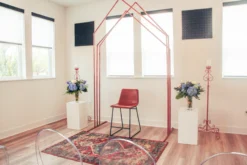 Copper arch with 3 peaks against a white wall with windows set up for a ceremony. Rug in front and florals on sides.