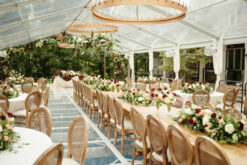 Wedding reception with a clear tent, a long row of farm tables and high end Louis chairs. Circular lighting strung from the ceiling