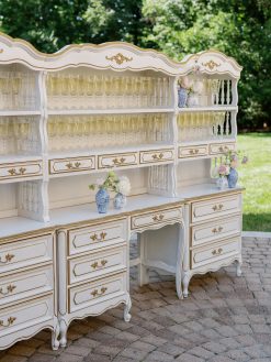 A grand, ornate white and gold bar hutch stands outdoors on a brick patio, ready for an event. Its shelves are neatly lined with rows of champagne flutes, and decorative blue and white vases filled with flowers are strategically placed on the lower surfaces.