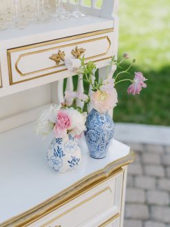 Two blue and white vases with flowers sit on a white bar cabinet with gold trim and ornate drawer pulls. The left vase holds white and pink flowers, while the right vase contains pink ranunculus and other delicate blooms.
