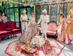 A wedding ceremony taking place on a red octagon patterned rug, with a couple in ornate traditional attire standing before a small altar, surrounded by guests in a conservatory venue.