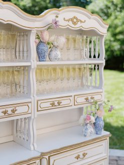 A vintage white bar hutch with gold trim and ornate details is filled with rows of champagne flutes. Two blue and white vases with flowers are placed on the bottom shelf, and two more on a higher shelf, adding a decorative touch.