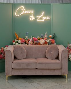 Pink velvet modern loveseat with a green backdrop and neon sign that says, 