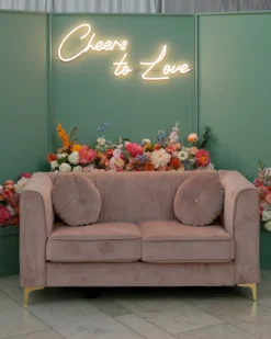 Pink velvet modern loveseat with a green backdrop and neon sign that says,