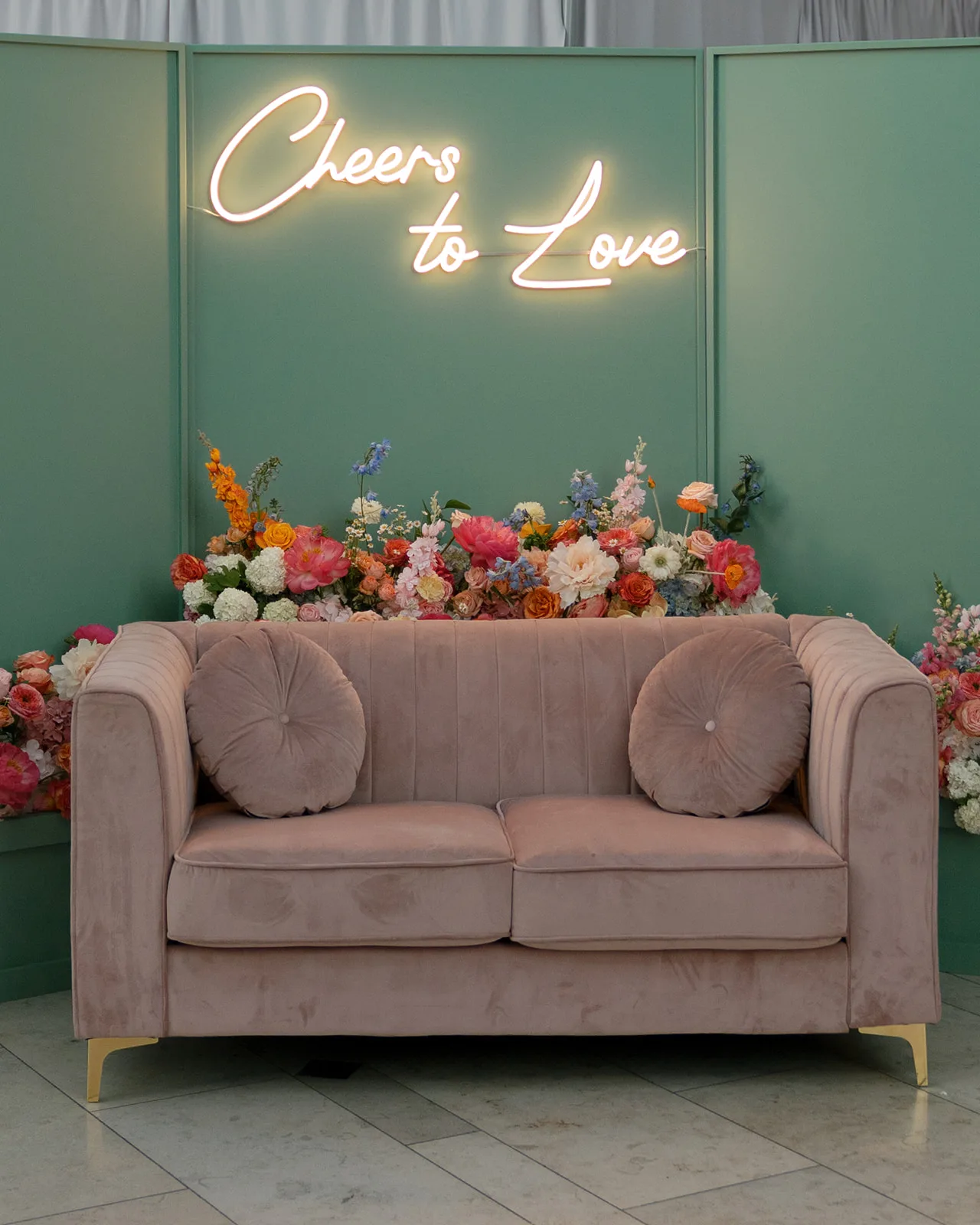 Loveseat for photobooth WEBP Pink velvet modern loveseat with a green backdrop and neon sign that says, "Cheers to Love" at a wedding reception