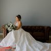 Bride in a white dress sitting on a brown leather sofa looking over the edge. Colorful rug in front of the sofa and her dress is spread out