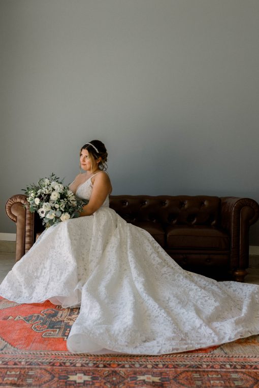 Bride in a white dress sitting on a brown leather sofa looking over the edge. Colorful rug in front of the sofa and her dress is spread out