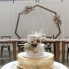 Cake table with a 2 tier white cake with florals on a large gold cakestand. Geometric backdrop with headtable and white barn doors.