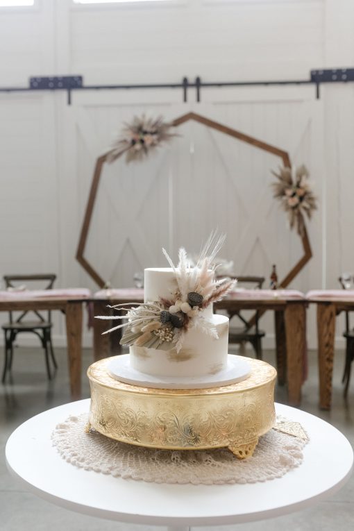 Cake table with a 2 tier white cake with florals on a large gold cakestand. Geometric backdrop with headtable and white barn doors.