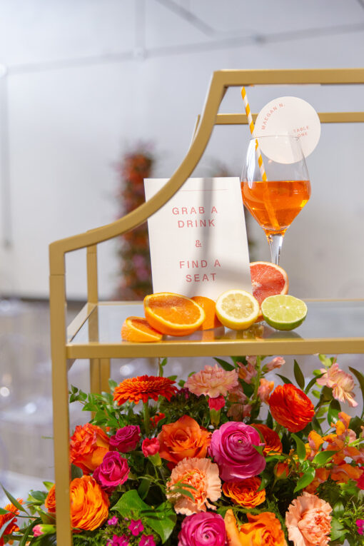 Close up on upper portion of gold shelf. Wine glass with bright orange welcome drink, sliced fruit and florals