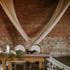 Sweetheart head table at a wedding reception with exposed brick wall and draping in the background. Wooden farmtable with two regal chairs with cream backs. Florals and tablescape in neutral clear glass and brown.