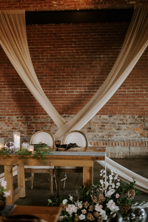 Sweetheart head table at a wedding reception with exposed brick wall and draping in the background. Wooden farmtable with two regal chairs with cream backs. Florals and tablescape in neutral clear glass and brown.