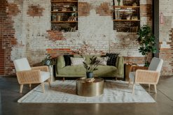A stylish event lounge setup featuring a moss green velvet sofa, two cane and white upholstered armchairs, and a hammered gold coffee table. The backdrop is an exposed brick wall with built-in bookshelves and a large plant.