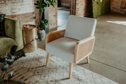 Close-up of a stylish accent chair featuring a light natural wood frame, woven cane side panels, and light gray upholstered seat and back cushions. Part of a lounge setup at an event.