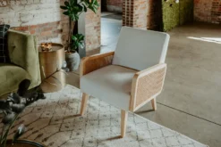 Close-up of a stylish accent chair featuring a light natural wood frame, woven cane side panels, and light gray upholstered seat and back cushions. Part of a lounge setup at an event.