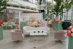 A beautifully decorated indoor event space with a white curved sofa, two light pink velvet chairs, and a glass-top side table. The area is adorned with vibrant floral arrangements and green plants. In the background, there are more flowers, tall green trees in large pots.