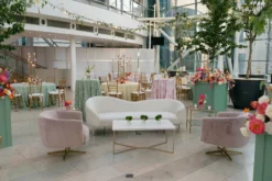 A beautifully decorated indoor event space with a white curved sofa, two light pink velvet chairs, and a glass-top side table. The area is adorned with vibrant floral arrangements and green plants. In the background, there are more flowers, tall green trees in large pots.