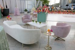 A beautifully decorated indoor event space with a white curved sofa, two light pink velvet chairs, and a glass-top side table. The area is adorned with vibrant floral arrangements and green plants. In the background, there are more flowers, tall green trees in large pots.
