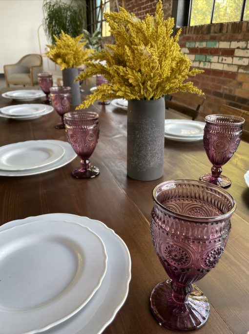 Wooden farmtable with white plates, plum matching goblets and goldenrod plant in a gray ceramic vase