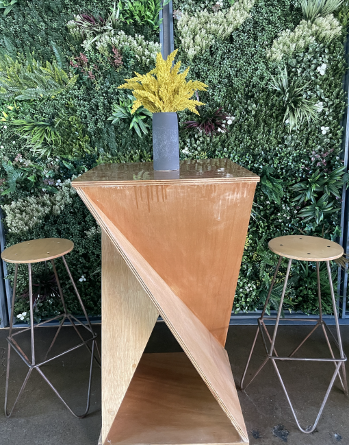 Wooden twist cocktail table with two metal legged stools. Goldenrod plant in a gray vase on the tabletop. Greenery wall background