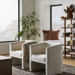 2 natural linen barrel chairs in a lounge with plant and bookshelf in background. Rug and coffee table corner in forground.
