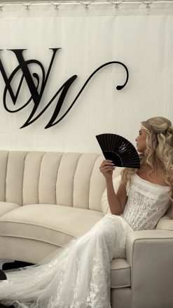A blond white woman in a white wedding dress with a black fan sitting on a velvet curved sectional.