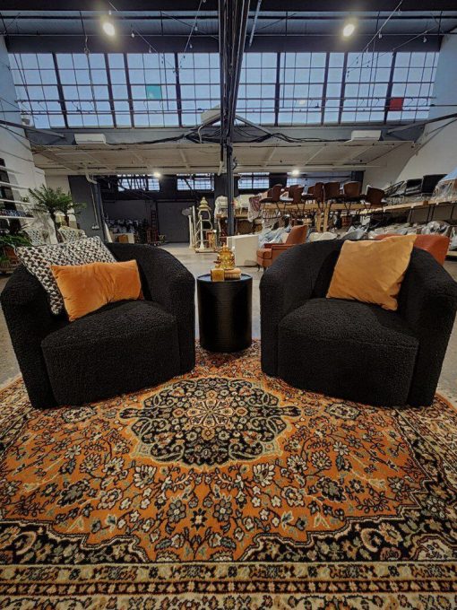 Two fuzzy black swivel chairs in a seated area with side table and a vintage orange rug. In an industrial space with high windows.