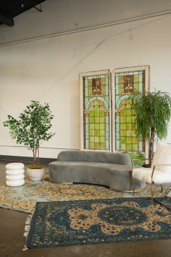 A stylish lounge seating area in a downtown Indianapolis event space featuring a curved grey velvet sofa, two vintage stained glass window backdrops, and plush layered oriental rugs. The setting is decorated with large indoor plants, a white fluted side table, and a white faux-fur armchair, creating a sophisticated atmosphere for a wedding reception or corporate event.