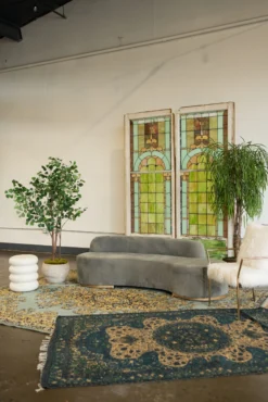 A stylish lounge seating area in a downtown Indianapolis event space featuring a curved grey velvet sofa, two vintage stained glass window backdrops, and plush layered oriental rugs. The setting is decorated with large indoor plants, a white fluted side table, and a white faux-fur armchair, creating a sophisticated atmosphere for a wedding reception or corporate event.