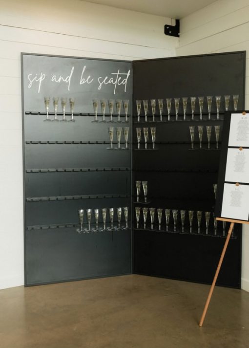 Black Champagne Wall with two panels. Each row holds 13 glasses, with 5 rows per panel. At the top of 1 panel, "sip and be seated" in script white letters.