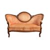 A vintage settee with a carved wooden frame and peach or coral velvet upholstery. The settee features a curved back with a central oval detail, serpentine arms, and cabriole legs. It has a white backdrop.