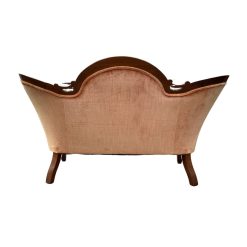 Back view of a vintage settee with a carved wooden frame and peach or coral velvet upholstery. The settee has a curved back with an oval center detail visible, and the wooden frame shows its ornate shaping. The back of the seat cushion and the tops of the curved legs are also visible against a white background.