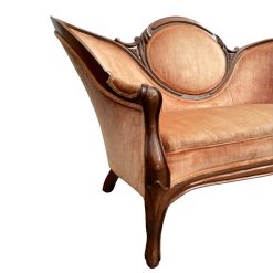 Close-up of a vintage settee with a carved wooden frame and peach or coral velvet upholstery. The image highlights the curved arm, the oval detail on the back, and the cabriole leg.