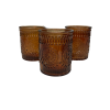 Three translucent, deep brown colored glass votive candle holders with an ornate, raised primrose or floral pattern on their surfaces. The votives are cylindrical