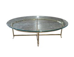 Travis Coffee Table for rent at Violet Vintage Indianapolis: A mid-century modern oval coffee table with a clear glass top, a gold-finished brass metal frame, and slim legs with decorative feet.