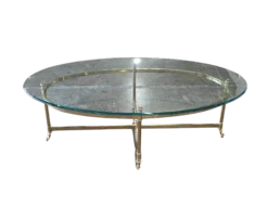 Travis Coffee Table for rent at Violet Vintage Indianapolis: A mid-century modern oval coffee table with a clear glass top, a gold-finished brass metal frame, and slim legs with decorative feet.