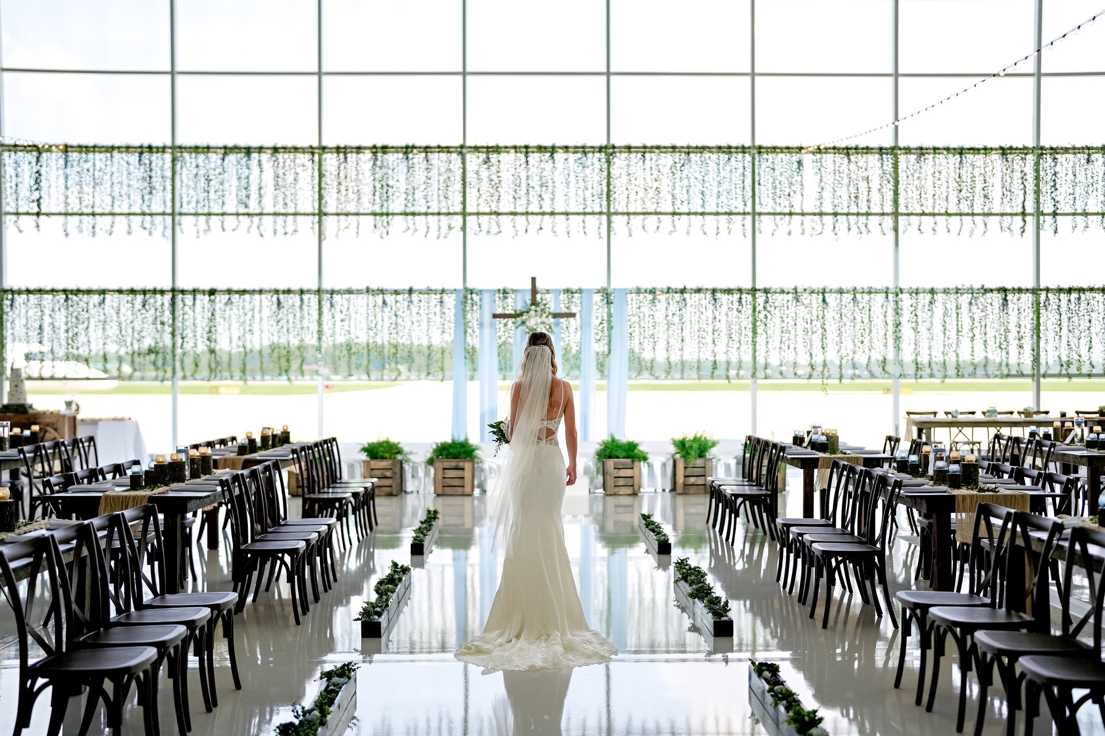 Bride walking through a ceremony aisle in an airport hangar with cross back chairs arranged beside reception tables.