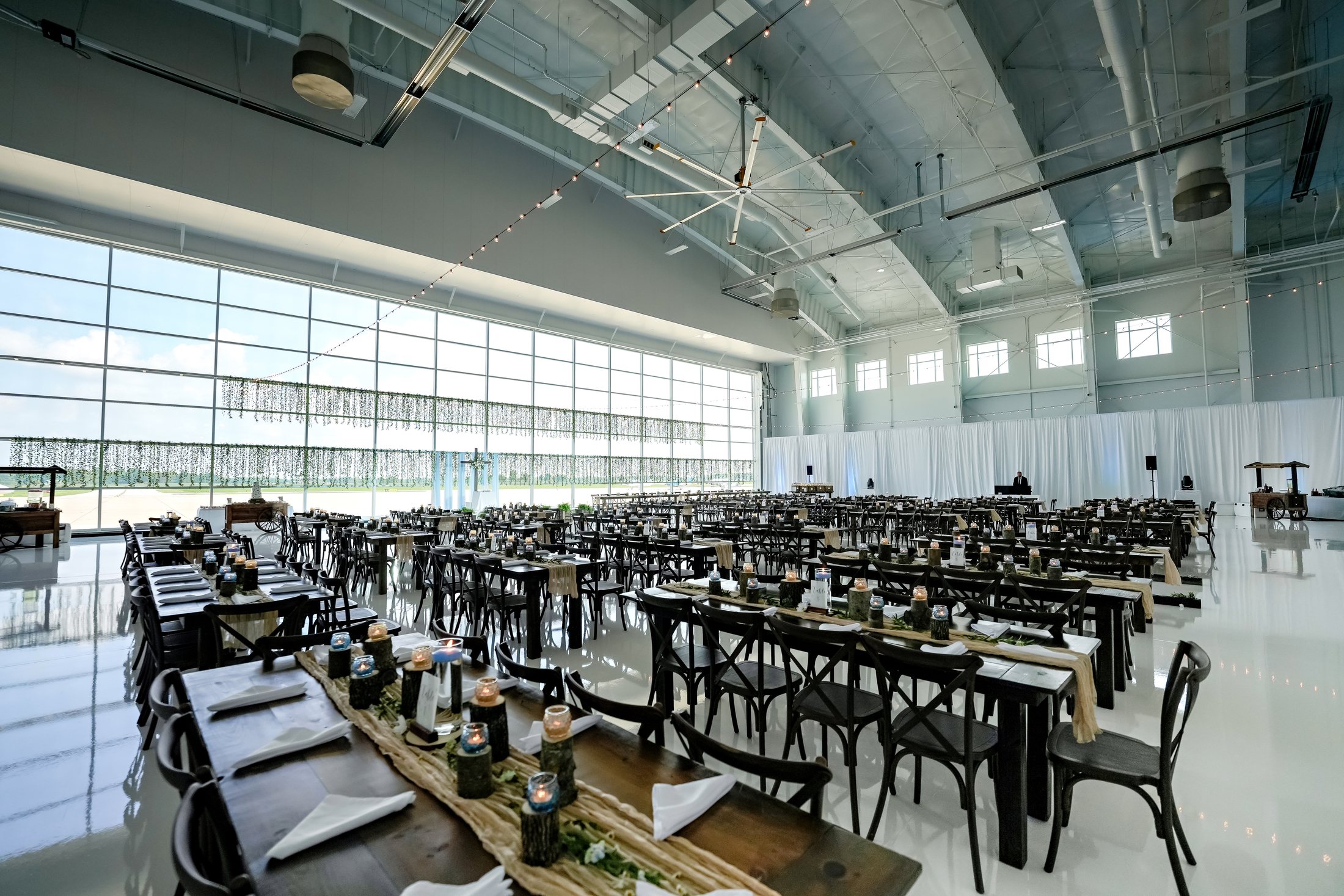 Rows of Dark Resin Vineyard Cross Back Chairs paired with Kingston Farmtables inside an airport hangar wedding reception.