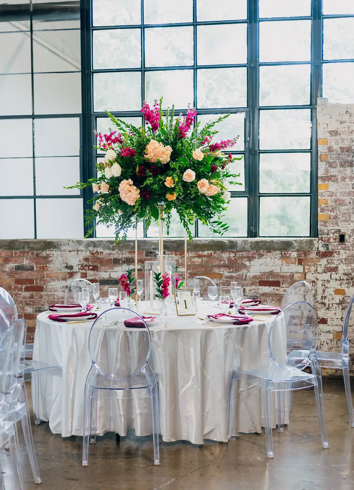 Round wedding reception table with clear Ghost Chairs, tall floral centerpiece, brick wall, and industrial windows at VV Showroom