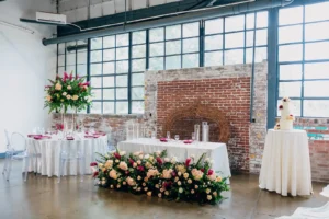 Wide view of Folake and Aloy's intimate wedding reception at VV Showroom in Indianapolis with Ghost Chairs, a floral head table, and cake display