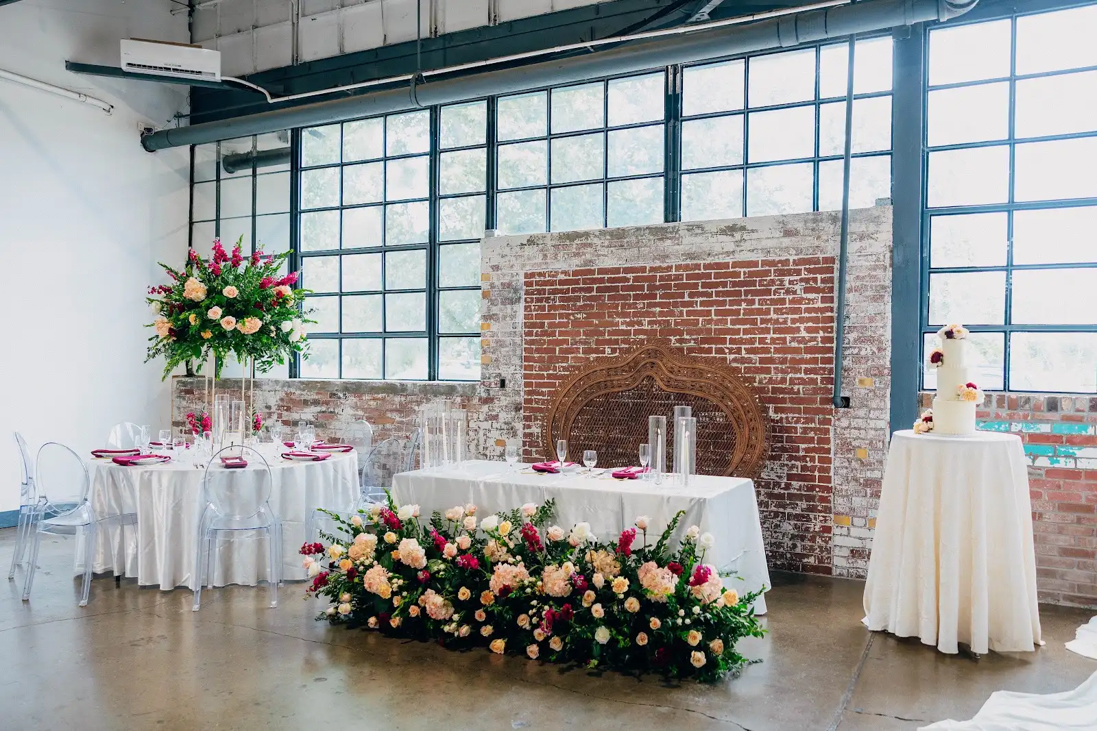 Wide view of Folake and Aloy's intimate wedding reception at VV Showroom in Indianapolis with brick walls, tall windows, Ghost Chairs, floral head table, and cake display
